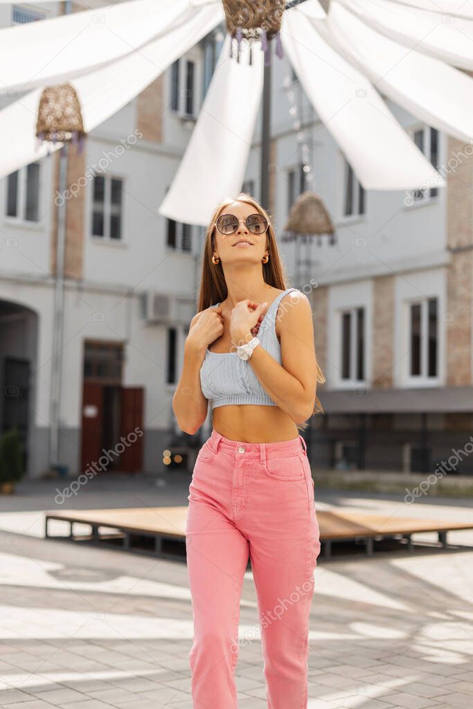 Happy beautiful young woman with vintage round sunglasses in bright summer outfit with blue top and pink pants walking in the city
