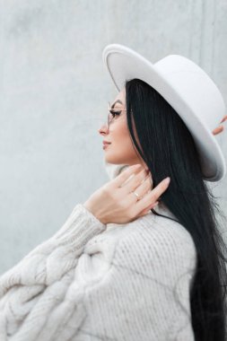 Gri bir arka planda şapkası ve gözlüğü olan, modaya uygun beyaz bir süveter giyen, uzun esmer, güzel bir kadın.