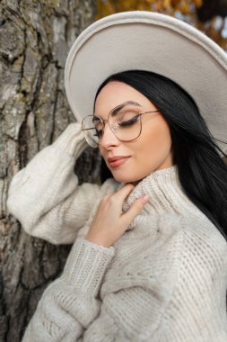Bir sonbahar parkında, bir ağacın yanında eski moda gözlüklü, şapkalı ve örgü süveterli güzel bir kadının sonbahar portresi.