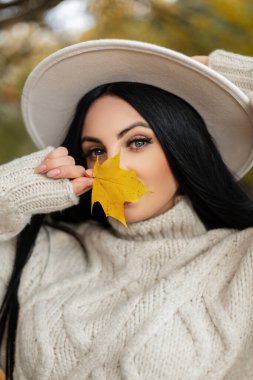 Parlak sarı bir sonbahar yaprağıyla şık, güzel bir genç kız ve kameraya bakıyor. Klasik örgü kazak ve şapkayla parkta yürüyen güzel bir kadın.