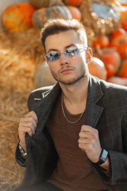 Paltolu ve sweatshirtlü, klasik güneş gözlüklü yakışıklı adam kırsalda bir balkabağının yanında oturuyor.