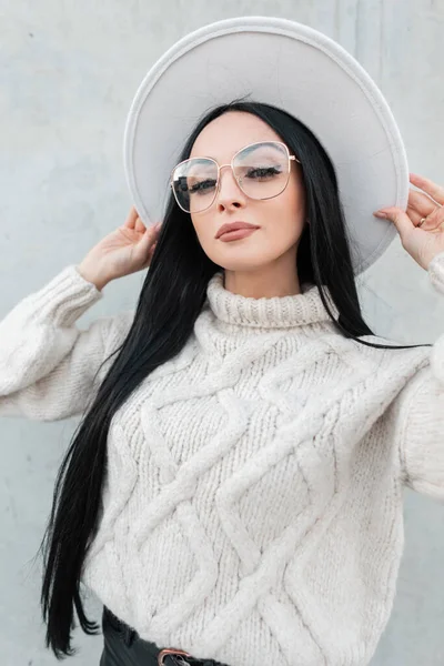 ファッショナブルな美しい女の子でスタイリッシュな眼鏡でヴィンテージニットセーターは 通りの灰色の壁の近くに帽子を着用し カメラを見て — ストック写真