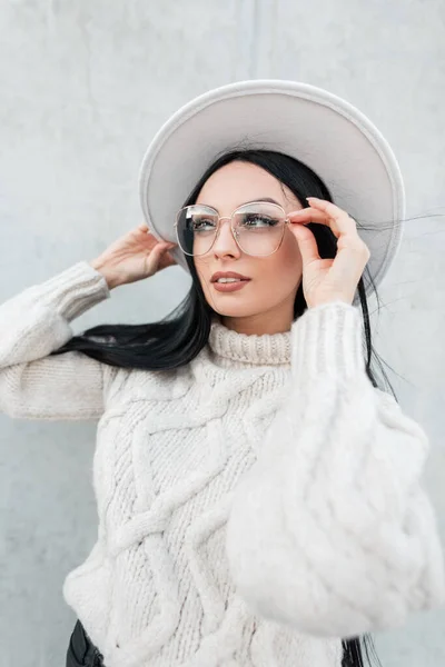 Female nice portrait of beautiful young with hat woman in fashion knitted sweater wears and puts on a stylish glasses on the street