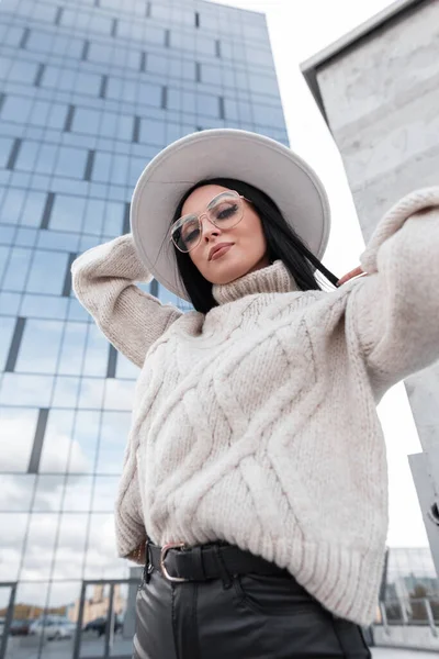 Urban female portrait of beautiful fashion woman model with stylish glasses in vintage cozy sweater and white hat travels in the city