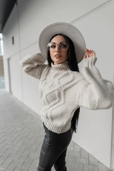 Fashion urban girl model with glasses in fashionable knitted sweater and hat walks on the street near a gray modern building