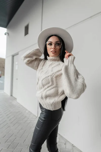 Pretty fashion Caucasian model girl with vintage glasses in trendy knit sweaters and white hat walks on the street near a gray wall. Female urban style outfit
