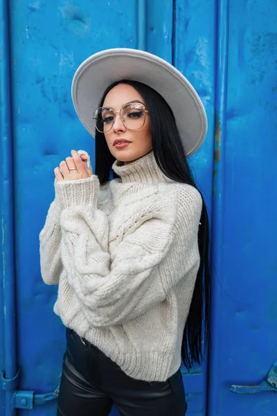 Fashion pretty woman with long hair and vintage glasses in trendy knitted sweater and hat poses near a colored blue metal background