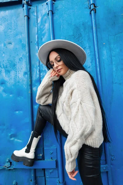 Cool fashion beautiful woman model with vintage glasses in white knitted sweater, hat, leather pants and shoes poses near a blue metal wall