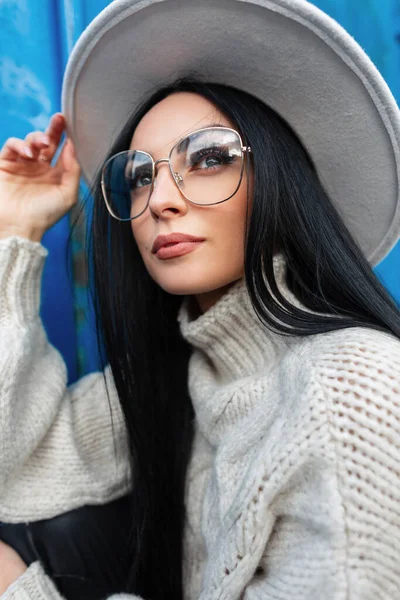 Urban female portrait of caucasian pretty woman with vintage glasses in fashion knitted sweater and hat stands and poses near a blue metal background on the street
