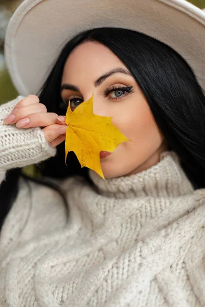 Autumn female portrait of young pretty woman with amazing blue eyes in vintage beige sweater and fashion hat covers her face with a bright yellow leaf and looks at the camera