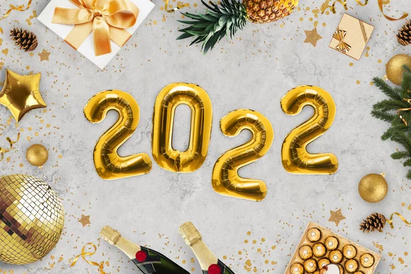 Luxury New Year 2022 party. Golden balloons 2022 lying on gray background with gold mirror ball, candy, champagne, gifts, pineapple, Christmas tree and candy, top view. Holiday celebrations.