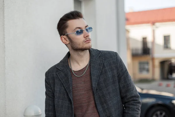 Handsome hipster man with vintage blue sunglasses in casual stylish coat walks in the city