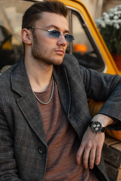 Trendy Handsome businessman hipster guy model with blue stylish sunglasses in a fashionable coat with a stylish luxury watch sits near a yellow retro car outdoor