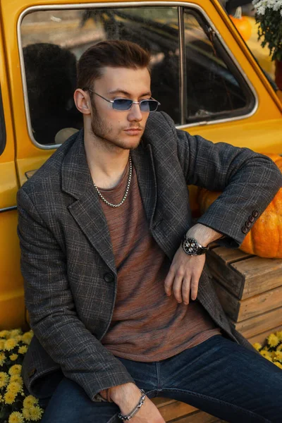 Fashionable hipster man with hairstyle and trendy vintage sunglasses in fashion coat, jeans and sweatshirt with luxury watch sits near yellow car countryside. Men\'s autumn style look clothes