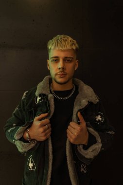 Havalı siyah ceketli, modaya uygun bir hippi, siyah bir duvarın yanında duruyor.