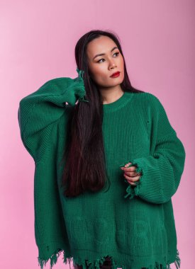 Moda yeşili yırtık kazaklı güzel Çinli manken kız stüdyoda uzun ve sağlıklı saçlarını pembe bir arka planda düzeltiyor.