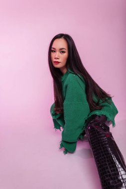 Moda kıyafetleri içinde, zarif yeşil kazağı ve deri pantolonu olan şık, güzel Koreli kız stüdyoda pembe bir arka planda oturuyor.