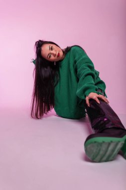 Pembe süveter, deri pantolon ve çizmelerle süslü, uzun saçlı güzel Asyalı manken pembe arka planda stüdyoda oturuyor.