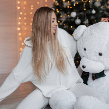 Güzel, genç, mutlu, sarı saçlı, modaya uygun beyaz elbiseli, kapüşonlu, oyuncak ayının ve Noel ağacının yanında oturan güzel bir kız.