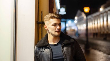 Şık, siyah deri ceketli, yakışıklı bir genç adamın gece portresi şehrin ışıklarla kaplı bir binasının yanında duruyor.