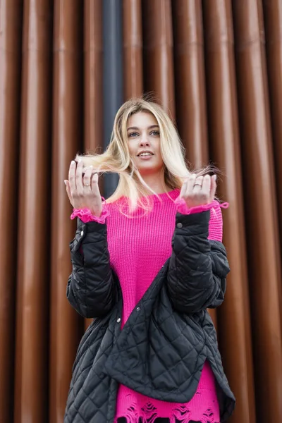 Beautiful happy fashionable woman with blonde hair in fashionable winter clothes with jacket and vintage pink sweater walking outside near a golden metal wall