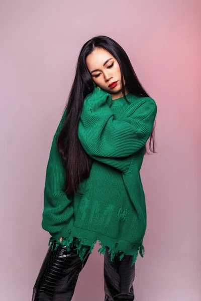 Fashion Asian pretty woman model with long hair in stylish knitted green sweater and leather pants stands in studio with pink light