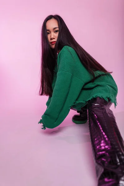 穿着时髦的撕破绿色毛衣 穿着时尚皮裤 脚踩着粉色彩灯的工作室里的一双靴子的美丽的年轻的亚洲时尚女孩 — 图库照片