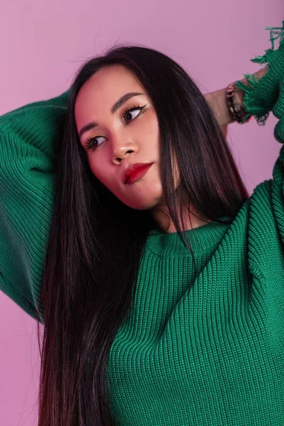 Beautiful young Asian female model with long hair and red lips in a green sweater on a pink background