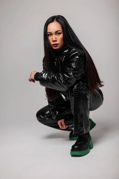 Stylish beautiful woman with makeup and red lips in fashionable black clothes with leather jacket, pants and shoes squatting and posing in the studio. Women\'s style black outfit