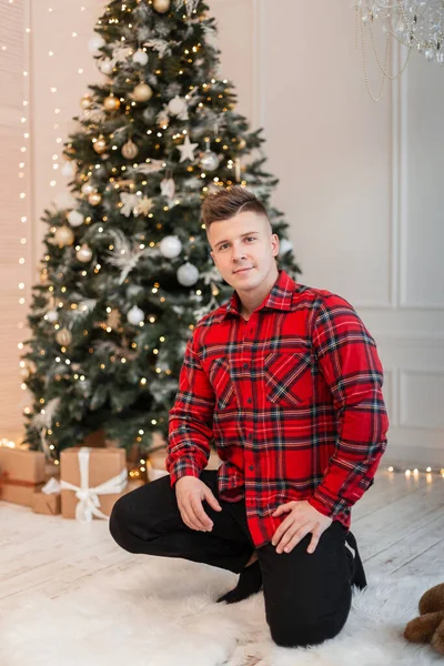 Hipster handsome man with hairstyle in a fancy red plaid shirt sits by the Christmas tree with lights at home