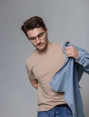 Saç stili ve klasik gözlüklü yakışıklı hippi çocuk gri arka plandaki stüdyoda kot gömlek giyiyor.