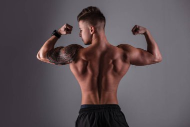 Kaslı arkası ve arkası kaslı yakışıklı vücut geliştirici stüdyoda koyu bir arka plana karşı kaslarını gösteriyor.