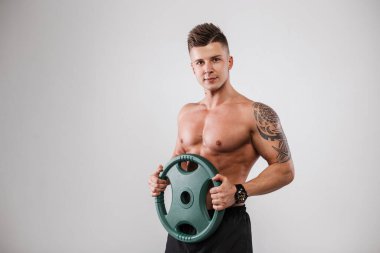 Dövmeli çıplak atletik gövdeye sahip güçlü kaslı atletik erkek modeli tarafsız gri arka planda stüdyoda ağırlığı tutuyor.