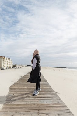 Kafasında eşarp, gözlüklü, yelekli ve spor ayakkabılı şık bir kız okyanus kıyısındaki tahta bir geçitte yürüyor. Kadınların bahar kıyafetleri.