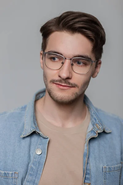 Studio male portrait of a handsome stylish guy with a beard and mustache with a haircut in fashionable denim clothes and vintage glasses on a gray background