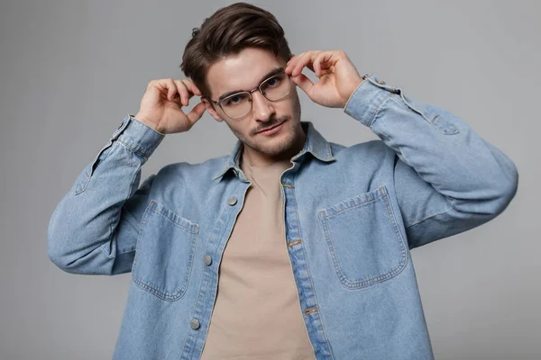 Stylish young businessman man with a haircut in trendy casual clothes with a denim shirt and T-shirt adjusts vintage glasses in the studio and looks at the camera