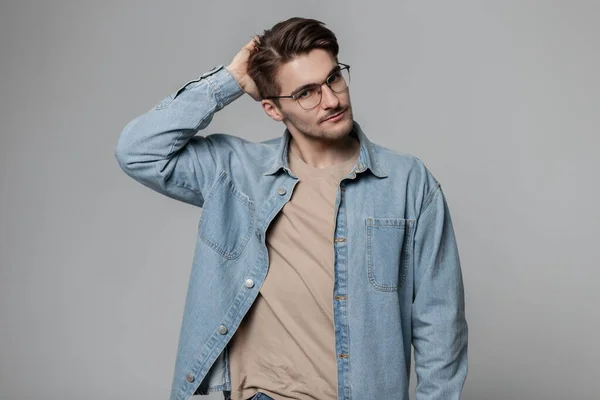 Cool handsome fashion student man with hairstyle in blu jeans shirt with vintage stylish glasses stands and poses on a gray background in studio