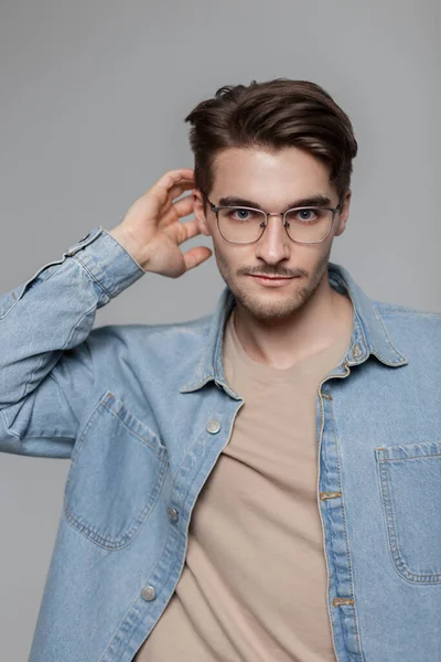 Fashion male portrait of handsome young american hipster man with vintage glasses and hairstyle in casual outfit with t-shirt and denim blue shirt poses and looks at the camera indoor
