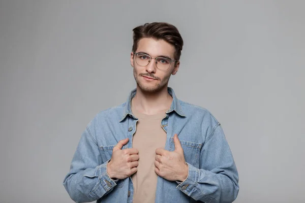 Fashionable American hipster man with vintage sunglasses, denim shirt and beige T-shirt on a gray background in the studio. Male portrait