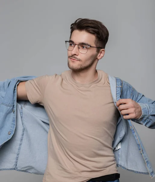 Fashionable American hipster model guy with hairstyle and vintage glasses in fashionable clothes with a beige T-shirt wearing a denim shirt on a gray background