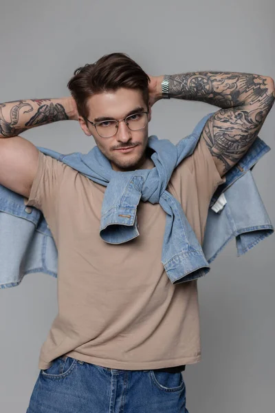 Trendy sexy hipster model man with vintage glasses wearing stylish clothes with a denim shirt and jeans and t-shirt poses with his hands behind his head in the studio and looks at the camera