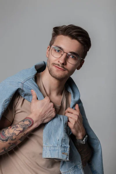 Fashionable handsome model guy with a mustache, beard and haircut in stylish clothes with a T-shirt and tied denim shirt on his shoulders with vintage glasses in the studio, looks at the camera.