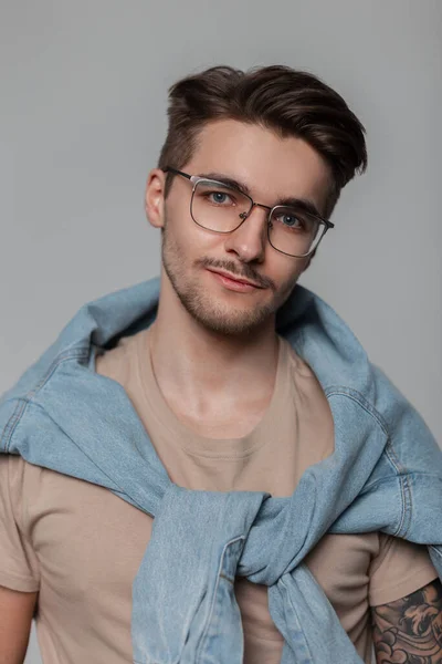 Stylish male portrait of a handsome young guy with hairstyle and vintage glasses in a fashion denim shirt looking at the camera against a gray background