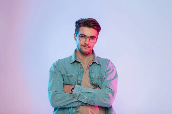 Fashionable colorful creative studio portrait of a handsome young guy with hair and vintage glasses wearing trendy denim clothes with a stylish denim shirt with pink and blue lights. Hipster man