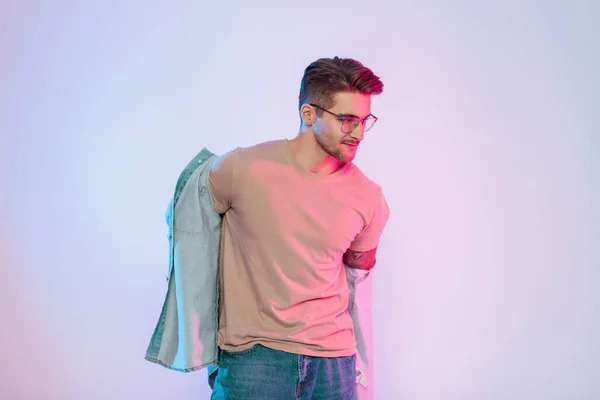 Handsome hipster man model with glasses in casual denim clothes wears jeans jacket in studio with creative colorful pink and blue lights