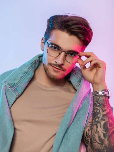 Cool fashion creative portrait of handsome young man model with hairstyle in trendy jeans outfit with tattoos on hand wears vintage fashionable glasses in studio with pink and blue lights