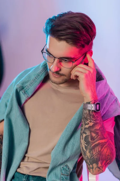 Fashionable handsome hipster guy with a haircut and vintage style glasses wearing trendy jeans and a retro watch sits in a studio with pink and blue lights. Trendy man with tattoo on hand