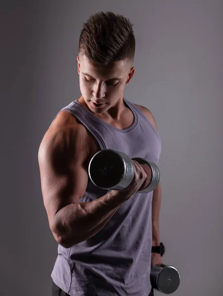 Handsome sporty man athlete with a muscular body pumps his biceps muscles and works out with dumbbells against a dark background in the studio