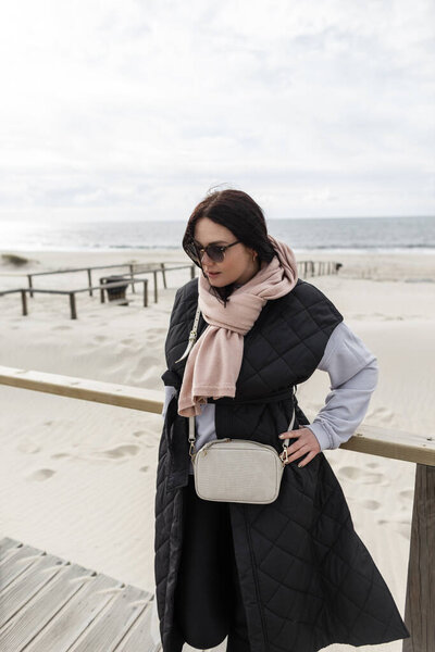 Beautiful young girl with vintage sunglasses in stylish spring clothes with a pink scarf, black vest and sweatshirt and purse walks on the beach by the ocean. Women's clothing collection