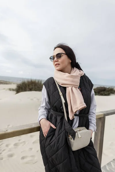 Beautiful fashionable woman with cool vintage sunglasses in a stylish black vest with scarf and sweatshirt walks on the beach by the ocean